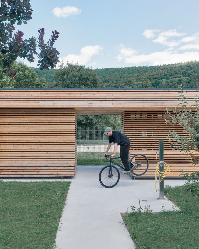 Cyclist riding under the projecting roof of a long, low larch-clad building