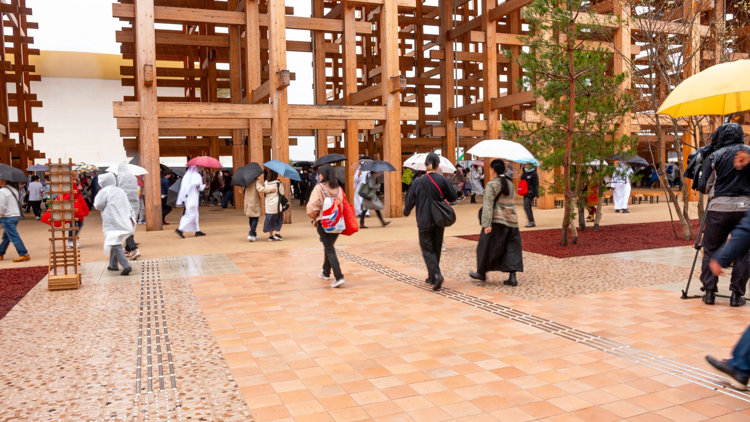 People with umbrellas walking on a paved square under a timber structure