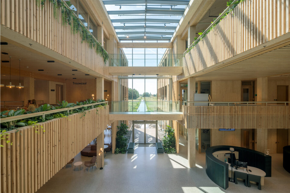 Atrium with timber slat balustrades, hanging plants, a glass roof and a tall window onto a ditch in the polder