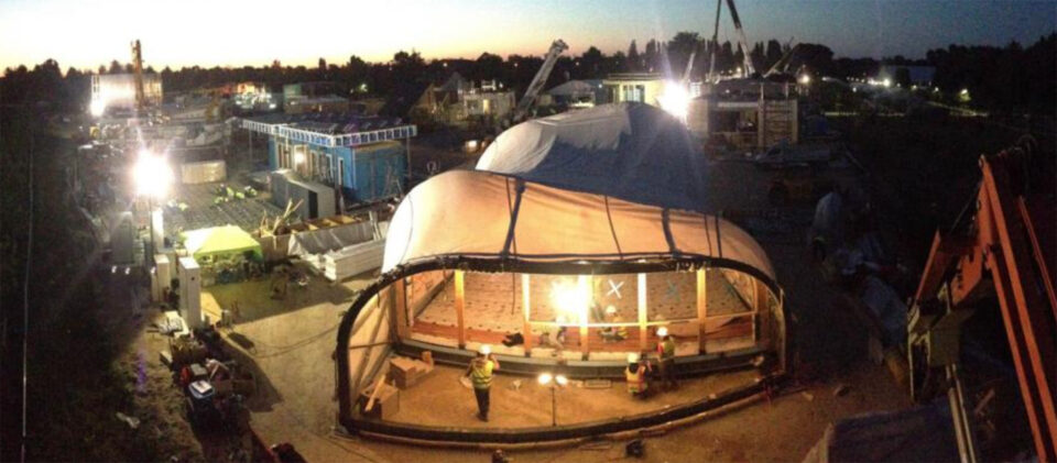 Student-built house with a curved white textile shell under construction at dusk, lit by floodlights