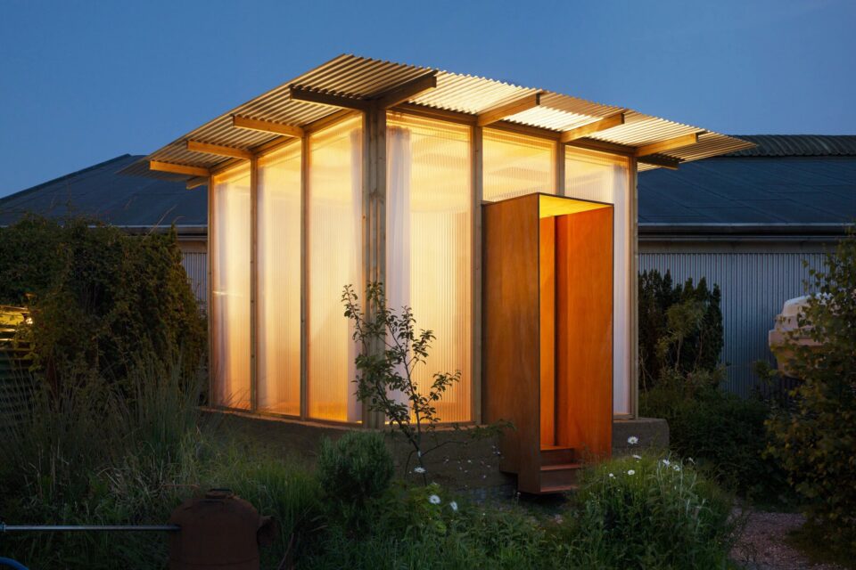 Small cabin with translucent walls glowing at dusk in a garden